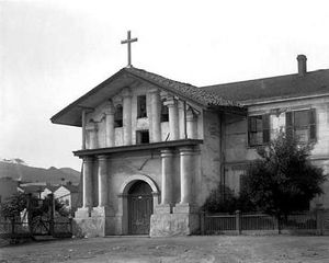 (PD) Photo: William Henry Jackson A view of Mission San Francisco de Asís (Mission Dolores) between 1880 and 1902. The stone building has an arched entryway, columns, a cross, and tile roof.