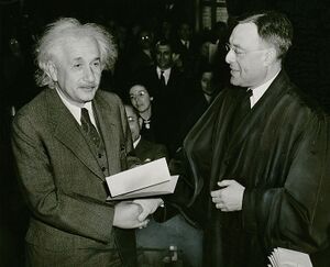 photograph of a white haired man on left (Albert Einstein) shaking hands with a man in a black robe.