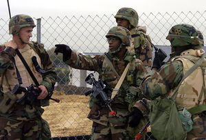 Picture of four soldiers outdoors in front of a fence; one soldier points to the left