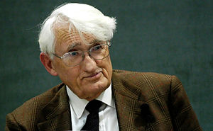 Picture of a man with white hair and glasses sitting behind a glass of water with a blackboard behind him.